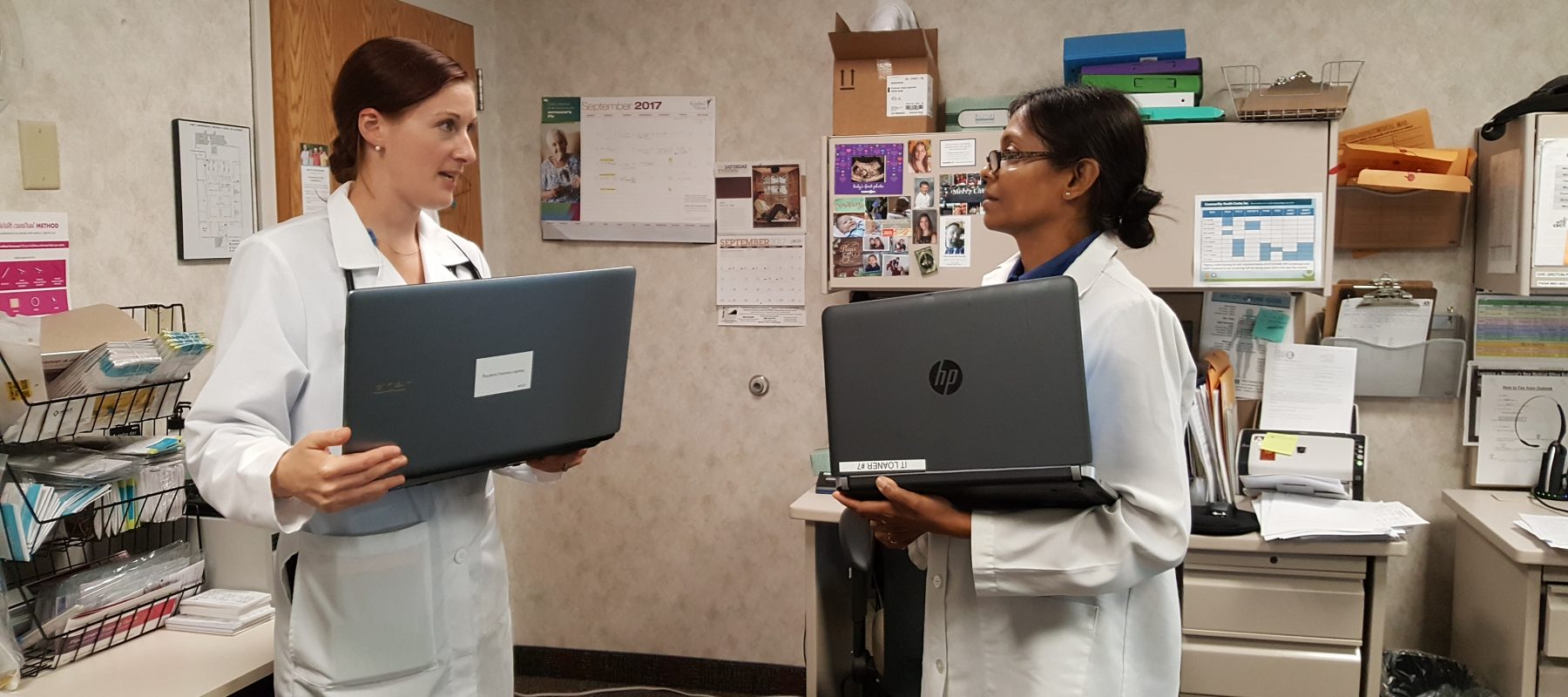 Two women in white coats talking to each other holding laptops in a doctors office